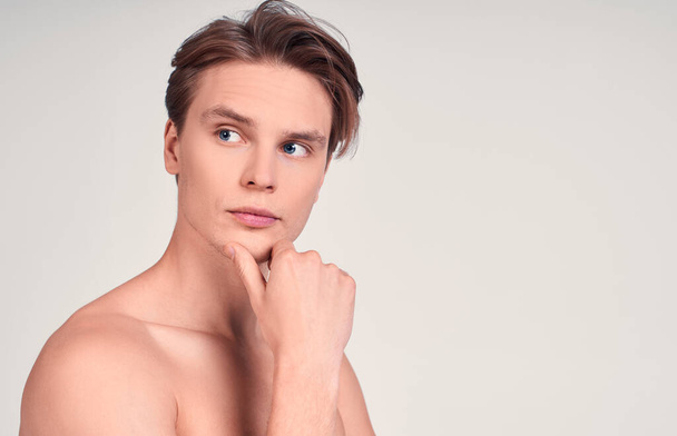 Close-up, beauty portrait of a Caucasian young man, with blue eyes and blond hair, standing against a white wall naked, touches his chin, looking to the side. The concept of beauty. Copy space. - Photo, image