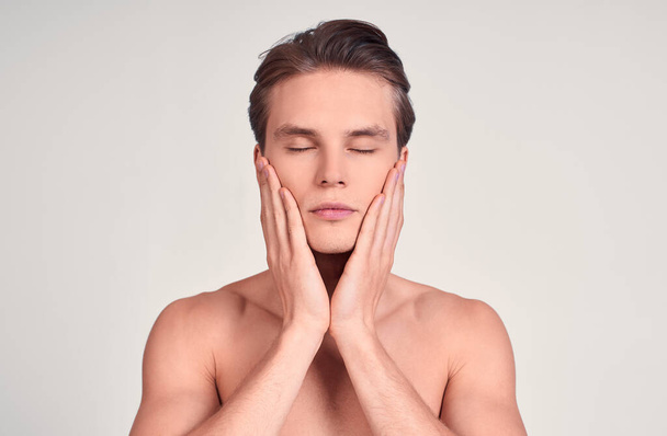 Young gorgeous Caucasian man with perfect hair and skin, shirtless, closed his eyes, touches his face with his hands, atmosphere of calm. Photo on a white background. - Photo, Image