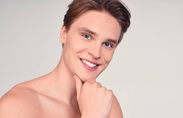 Portert of a charming cute attractive young guy, naked, with blond hair and blue eyes, with healthy skin, holds his hand near his chin, smiles and looks at the camera. - Photo, Image
