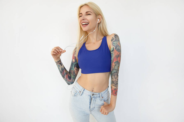 Good looking young tattooed long haired woman dressed in blue shirt and jeans smiling happily while listening to music in her earphones, isolated over white background - Photo, image