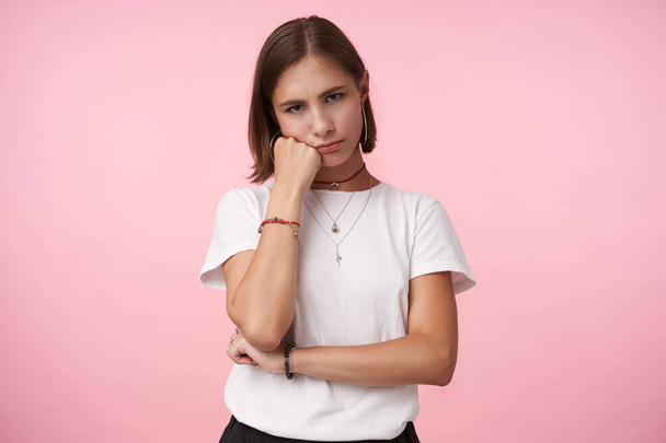 Indoor shot of upsed young short haired lady with casual hairstyle leaning her cheek on raised hand and looking sadly at camera, posing over pink background - Photo, Image