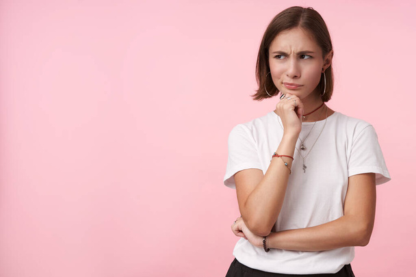 Bewildered young pretty dark haired woman dressed in basic white t-shirt twisting her mouth and frowning eyebrows while looking pensively aisde, posing over pink background - Foto, Bild