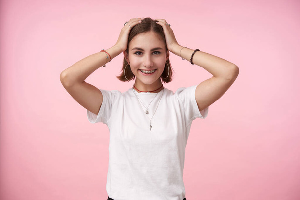 Inddoor photo of young charming dark haired woman keeping her hands on head while looking positively at camera with broad smile, standing over pink background - Fotoğraf, Görsel
