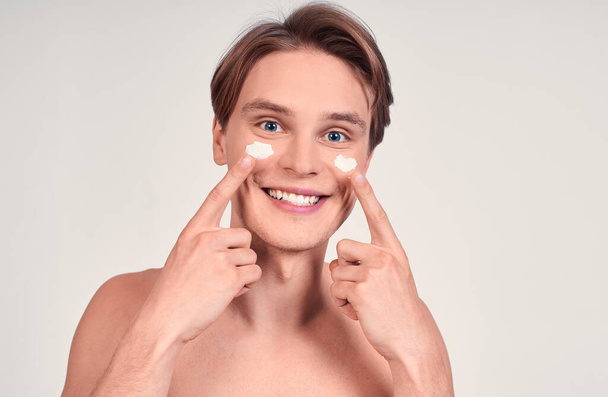 Photo of joyful half-naked man with facial cream smiling and looking at camera isolated over white background. Beauty procedure and skin care. - Фото, зображення