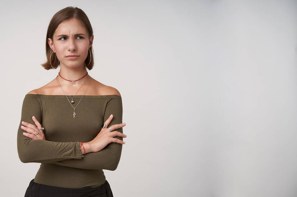 Bewildered young dark haired lady with short haircut folding hands on her chest while looking confusedly aside, dressed in olive blouse while posing over white background - Foto, Bild