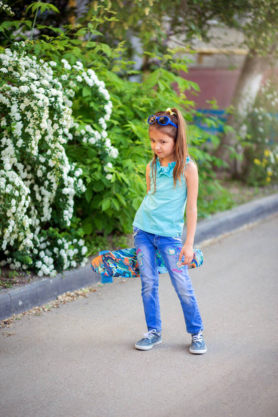Pequeña chica guay en jeans se para en el pavimento y sostiene un patín en sus manos. Deportes infantiles en casa
 - Foto, imagen