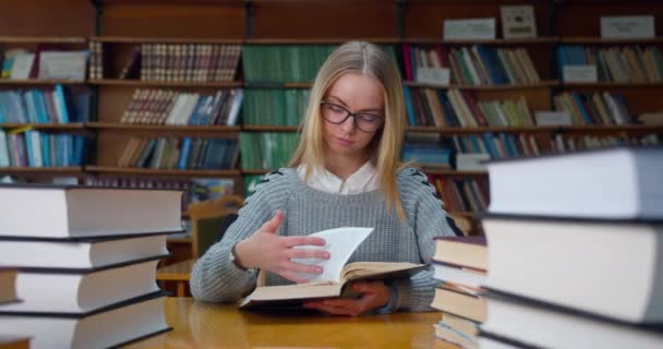 Das Mädchen studiert zwischen vielen Büchern im Lesesaal der Bibliothek. Schöne Mädchen mit Brille. 4K - Filmmaterial, Video