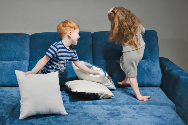 A little boy and a little girl with red hair are fighting with pillows. Brother and sister play with pillows on a blue sofa. - Foto, imagen