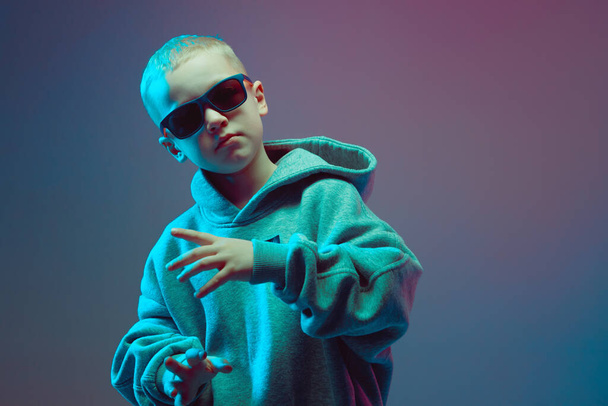 Portrait of a cool boy child in a rap image, stylishly posing in a hoodie, sunglasses and on a neon background. - Foto, afbeelding