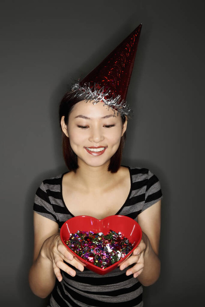 Woman with party hat holding a heart-shaped bowl of confetti - Foto, Bild