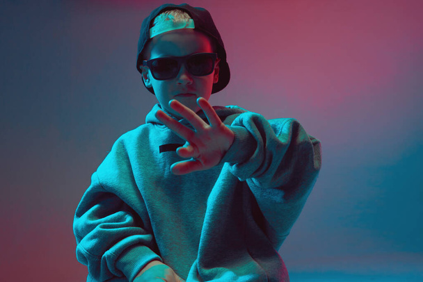 Portrait of a cool boy child in a rap image, stylishly posing in a hoodie, sunglasses and a cap on a neon background. - Фото, зображення
