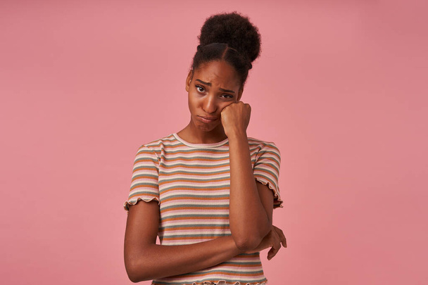 Unhappy young brown-eyed curly female with dark skin holding her face with raised hand while looking sadly at camera, going to cry while posing over pink background - Photo, Image