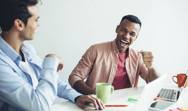 Porträt eines überglücklichen afroamerikanischen männlichen Programmierers, der das Ende des Startups mit der Erstellung von Webseiten feiert, die am Schreibtisch mit einem Kollegen sitzen und in die Kamera lächeln, Konzept zufriedener Menschen - Foto, Bild