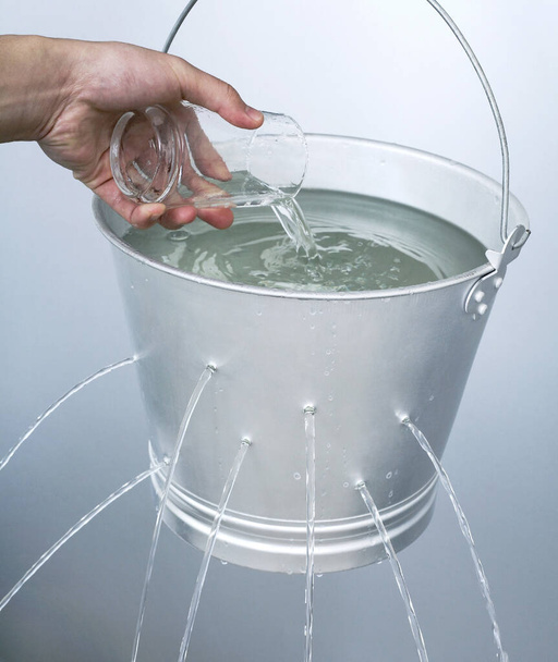 Hand pouring water from a glass into a leaking pail - Fotografie, Obrázek