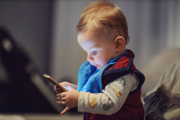 Portrait of cute blond toddler sitting in mother's lap, holding smart phone and touching screen. He is a big boy now and he have to learn how to post a picture on social media. - Photo, Image