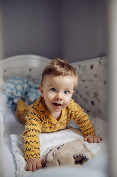 Adorable curious smiling blond baby boy with beautiful blue eyes lying on stomach in his crib and looking at camera. Morning time. - Fotografie, Obrázek