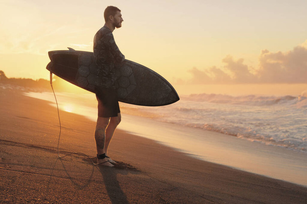 Surfing. Surfer With Surfboard Standing On Ocean Beach At Beautiful Sunrise In Bali. Man Silhouette On Attractive Sea And Sky Background. - 写真・画像