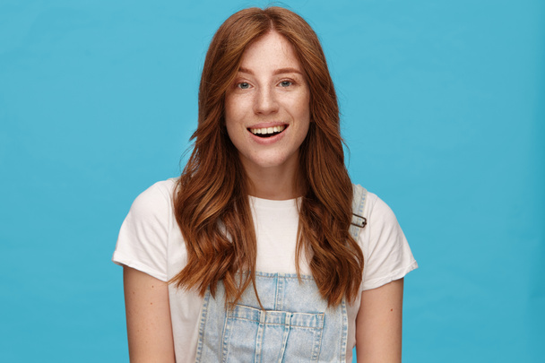 Joyful young charming redhead woman with natural makeup looking gladly at camera with broad sincere smile, being in nice mood while posing over blue background - Photo, Image