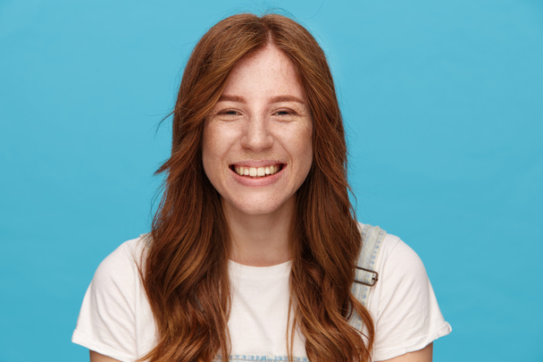 Close-up of young attractive green-eyed redhead woman showing her pleasant emotions and looking at camera with charming smile, isolated over blue background - Photo, Image
