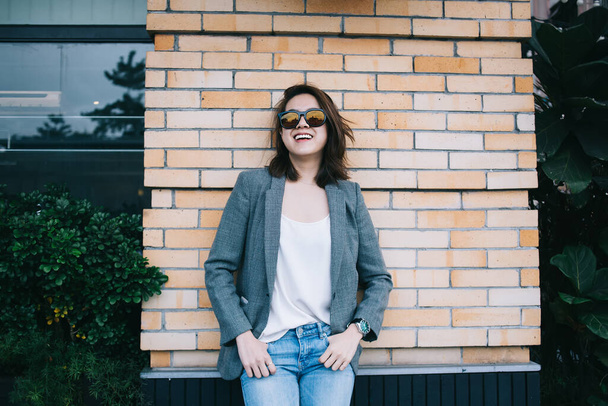 Laughing happy attractive brunette in sunglasses and trendy gray jacket with denim standing with hands in pocket and smiling against brick wall  - Fotografie, Obrázek