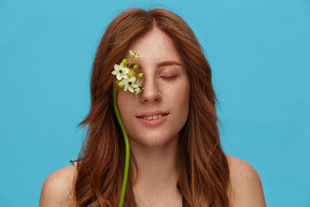 Крупним планом приємно виглядає молода позитивна руда леді з білими квітами на її обличчі, ніжно посміхаючись, стоячи над синім тлом із закритими очима
 - Фото, зображення