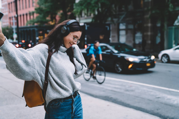 Cheerful casual woman in sweater and glasses walking on street and dancing while listening to music with headphones on New York street - Zdjęcie, obraz