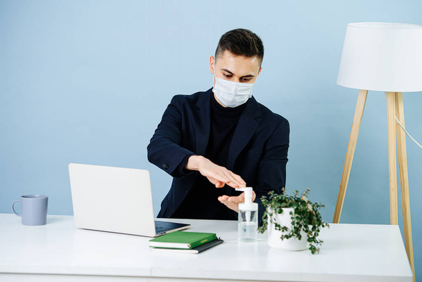 Masked man working in an office during covid-19 pandemic. He is disinfecting his hands before touching anything on his work desk. - Photo, image