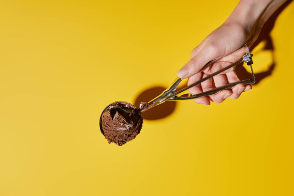 partial view of woman holding fresh chocolate ice cream ball in scoop on yellow background - Foto, imagen