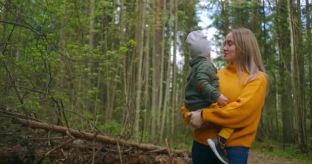 In slow motion, a mother and son walk through the Park in sweaters. A loving mother walks with her son in her arms in a pine forest. Mothers day. Caucasian woman with a funny boy 2 years old. - Footage, Video