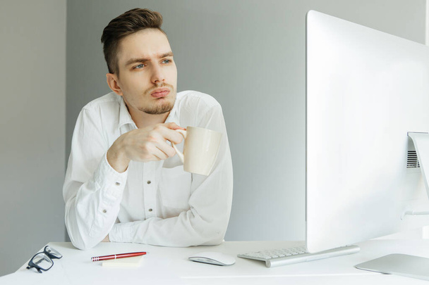 Young man in white shirt with cup working at the desktop with a computer in office. Worker on grey background - Photo, image