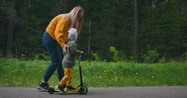 Een jonge moeder en haar zoon leren op een scooter te rijden. Moederdag. In een dennenbos op de weg gelukkig twee mensen een vrouw en een kleine jongen samen op een scooter. Hulp en opvoeding van het kind. - Video