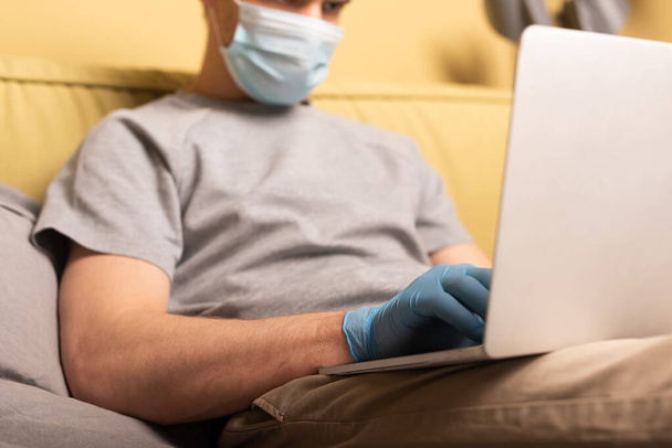Selective focus of man in medical mask and latex glove using laptop in bedroom - Fotó, kép