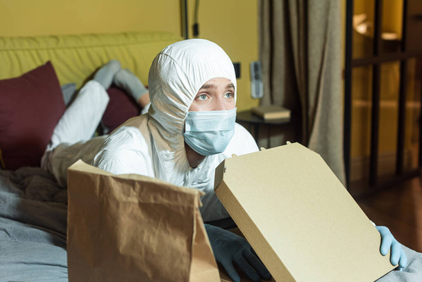 Enfoque selectivo del hombre en máscara médica y traje de materiales peligrosos viendo la televisión en la cama cerca del paquete y la caja de pizza
  - Foto, imagen