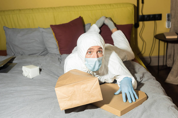 Hombre con traje de materiales peligrosos y máscara médica acostado cerca de paquetes y caja de pizza en la cama
  - Foto, Imagen