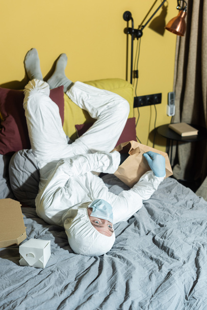 Homme en masque médical, gants en latex et combinaison Hazmat près des paquets et boîte à pizza sur le lit
  - Photo, image