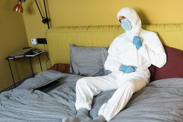 Selective focus of man in hazmat suit, medical mask and latex gloves sitting near laptop on bed  - Fotó, kép