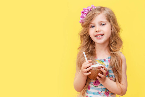 A cheerful little blonde girl drinks a coconut cocktail. A child in a swimsuit with a flower in her hair, summer vacation on the kunikulah.  - Foto, Imagem