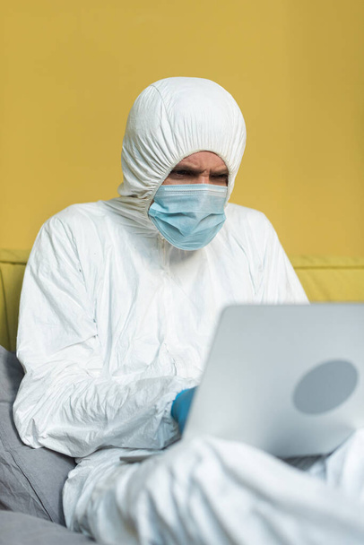 Foco seletivo do homem em terno hazmat e máscara médica usando laptop na cama
  - Foto, Imagem