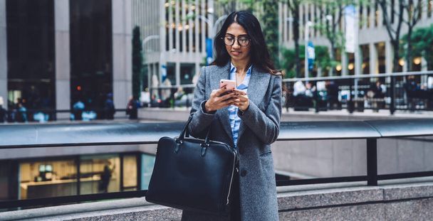 Contemporary trendy ethnic lady in glasses and coat holding leather briefcase and texting on mobile phone on background of New York city - Фото, изображение