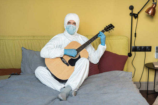 KYIV, UKRAINE - 24 Nisan 2020: Tehlikeli madde giysisi ve tıbbi maskeli bir adam akustik gitar tutuyor ve kanepede kameraya bakıyor  - Fotoğraf, Görsel