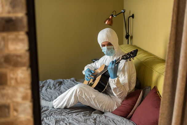 Επιλεκτική εστίαση του ανθρώπου με στολή Hazmat και γάντια λατέξ παίζοντας ακουστική κιθάρα στην κρεβατοκάμαρα - Φωτογραφία, εικόνα