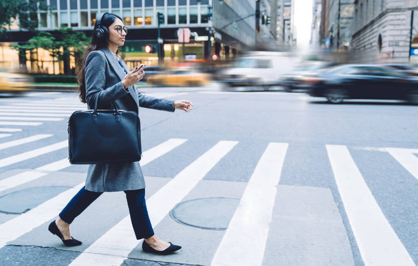 Side view of concentrated young brunette ethnic woman in earphones round spectacles gray coat with black handbag and smartphone crossing New York busy road - Valokuva, kuva