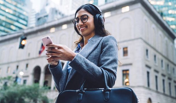 From above content young brunette ethnic woman in earphones round spectacles black handbag and gray coat tapping on smartphone on New York street on urban blurred background - Foto, Imagen