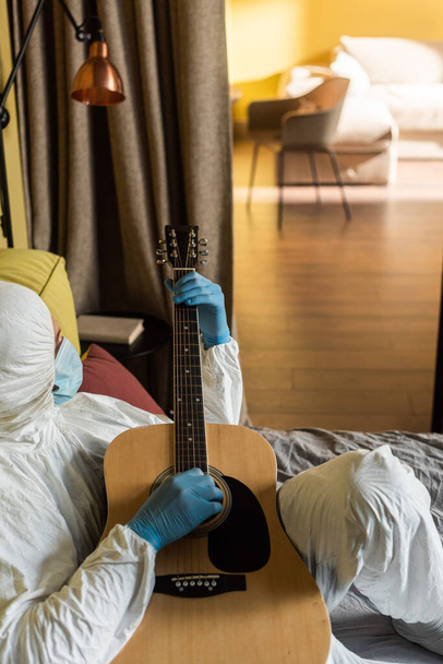Πλευρική άποψη του ανθρώπου με στολή Hazmat, ιατρική μάσκα και γάντια λατέξ που παίζουν ακουστική κιθάρα στο κρεβάτι  - Φωτογραφία, εικόνα