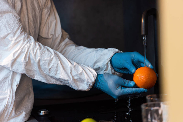 Ausgeschnittene Ansicht eines Mannes mit Latexhandschuhen und Schutzanzug, der Orange in der Küche wäscht  - Foto, Bild