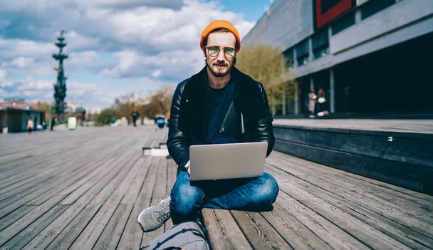 Portrait of stylish male it professional working remotely with laptop device on fresh air in city, trendy man in eyewear looking at camera while resting on publicity area with netbook for communicate - Foto, Imagem