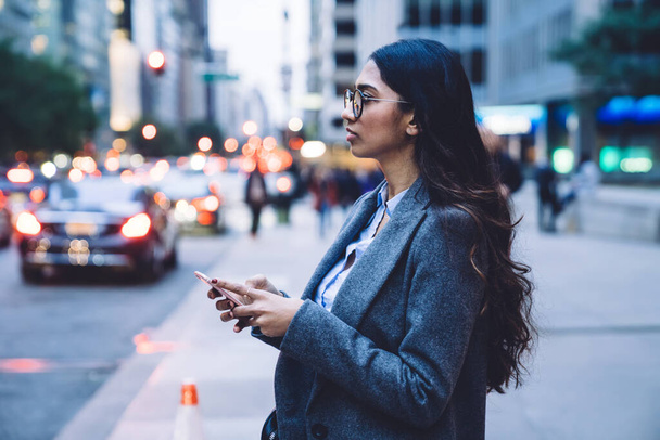 Side view of attractive ethnic woman in gray coat standing on roadside with smartphone in hands and thinking in New York City - Fotografie, Obrázek
