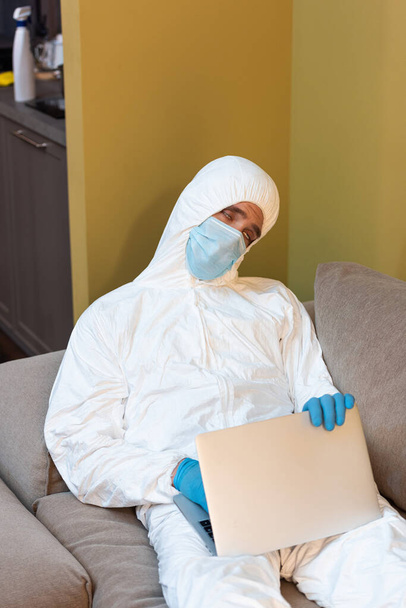 Tired man in medical mask, latex gloves and hazmat suit holding laptop on couch  - Foto, imagen