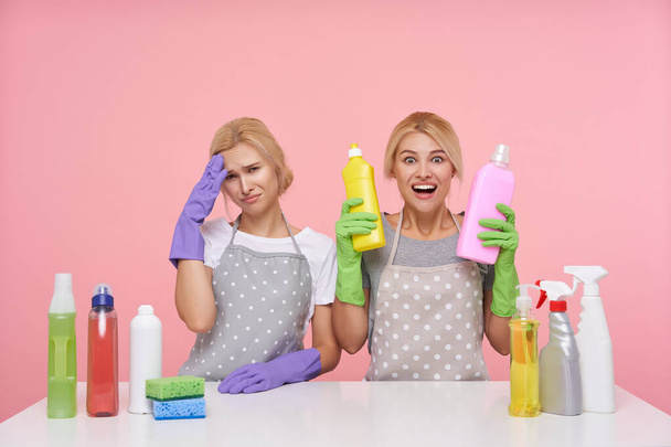 Unzufriedene junge weißköpfige Putzfrau blickt traurig in die Kamera, während ihre aufgeregte blonde Freundin freudig ihre Hände mit Waschmitteln hebt, isoliert vor rosa Hintergrund - Foto, Bild