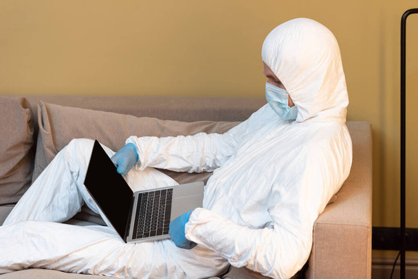 Zijaanzicht van de mens in medisch masker, latex handschoenen en hazmat pak met laptop op de bank in de woonkamer - Foto, afbeelding